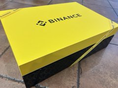 The image features a yellow box with "Binance" written on it, placed on a tiled floor. The box has a black border around its edges, giving it an elegant appearance. It appears that someone has just received this item or is preparing to send it out.