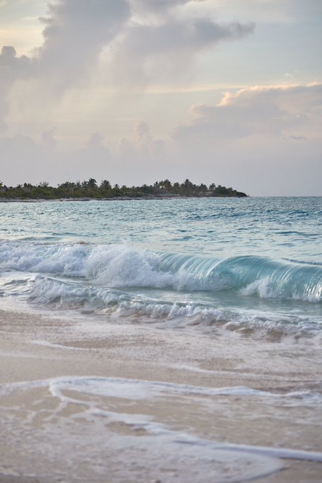A serene beach scene with gentle turquoise waves rolling onto white sand, with a small island covered in palm trees visible in the distance under a cloudy sky.