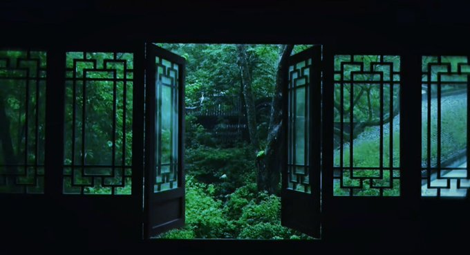 A view through open decorative wooden lattice windows looking out onto a lush green garden with trees.