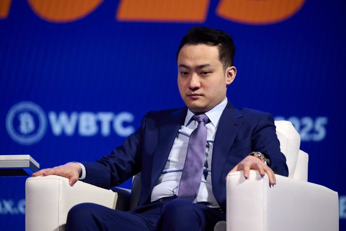 The image shows a man sitting in an office chair with his hands resting on his lap. He appears to be dressed in a suit and tie, suggesting that he might be attending a formal event or working at a professional setting. The background of the image features a blue wall with white text, which could possibly be related to the post about TRON's growth and expansion.