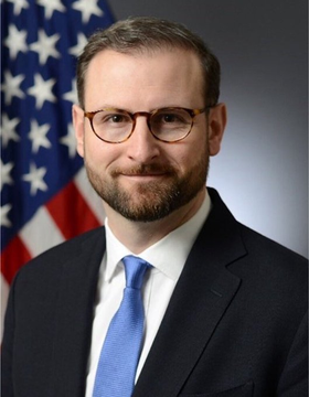 The image is an official portrait of a man with glasses and a beard, wearing a dark suit and blue tie, standing in front of an American flag. Based on the accompanying post, this appears to be the White House official Patrick Witt referenced in the caption about the CLARITY Act.