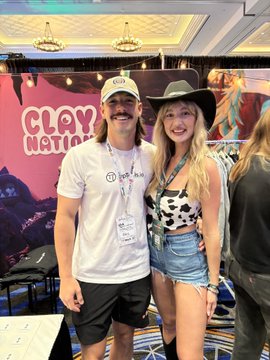 The image features two people posing for a picture, with one person wearing a cowboy hat and the other wearing a white shirt. They are standing next to each other against a pink wall adorned with a cartoon character design. The background includes several chairs arranged in various positions around them. A sign is also visible on the wall that reads "Clay Nation".
