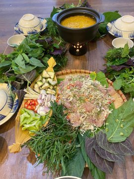 An overhead view of a table set with a pot of yellow soup and platters of fresh herbs, sliced vegetables, and raw fish/meat for a shared meal.