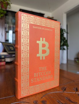 The image features a wooden table displaying three leatherbound books titled "The Bitcoin Standard" by Saifedean Amour. The books are arranged in a row with the largest book being positioned at the front, while the other two books are placed behind it. The books appear to be of interest to collectors or enthusiasts of cryptocurrencies like Bitcoin.
