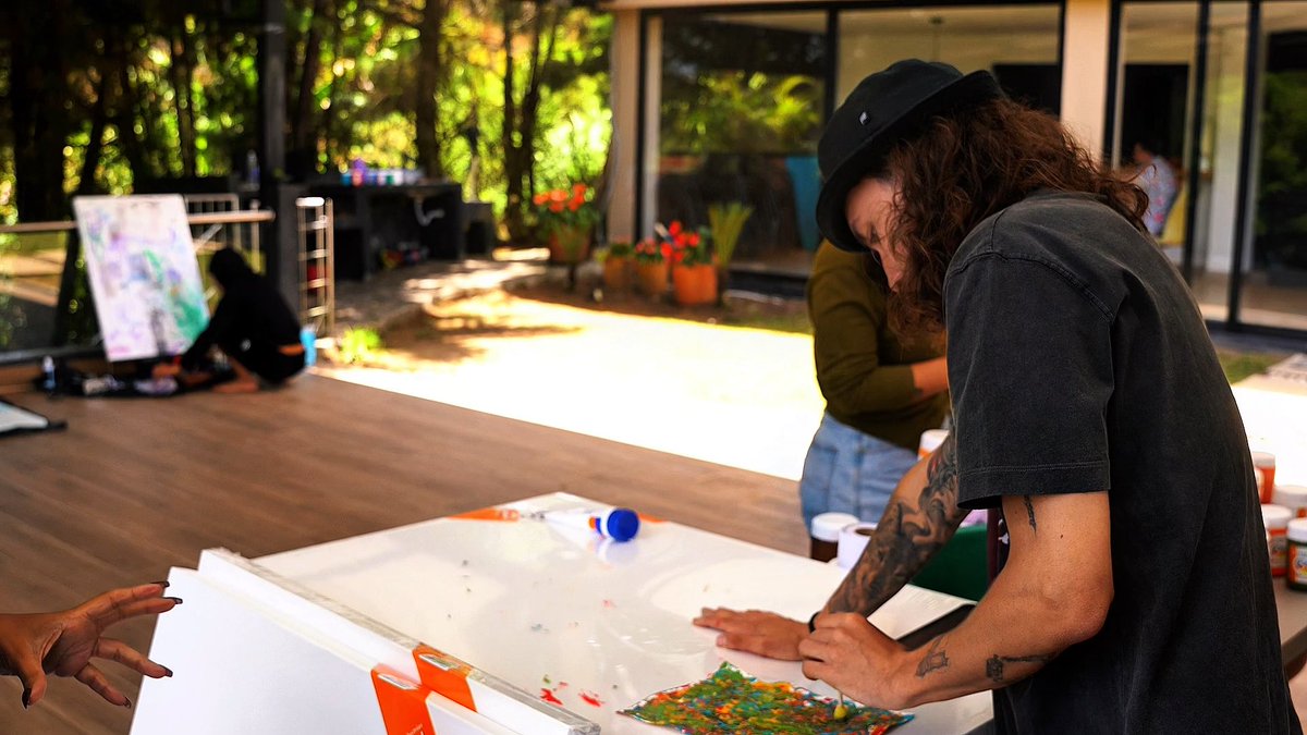 A candid outdoor scene at an art retreat showing people creating art, with one person in the foreground with tattoos and a black hat working on a colorful painting.