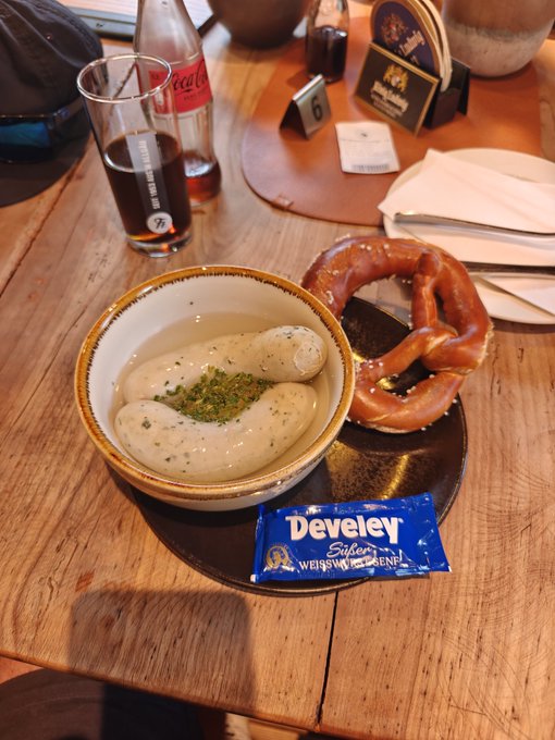A bowl of Bavarian-style white sausages served with a soft pretzel on a wooden table, accompanied by a Develey Süß-Senf sweet mustard packet and a glass of dark beer.