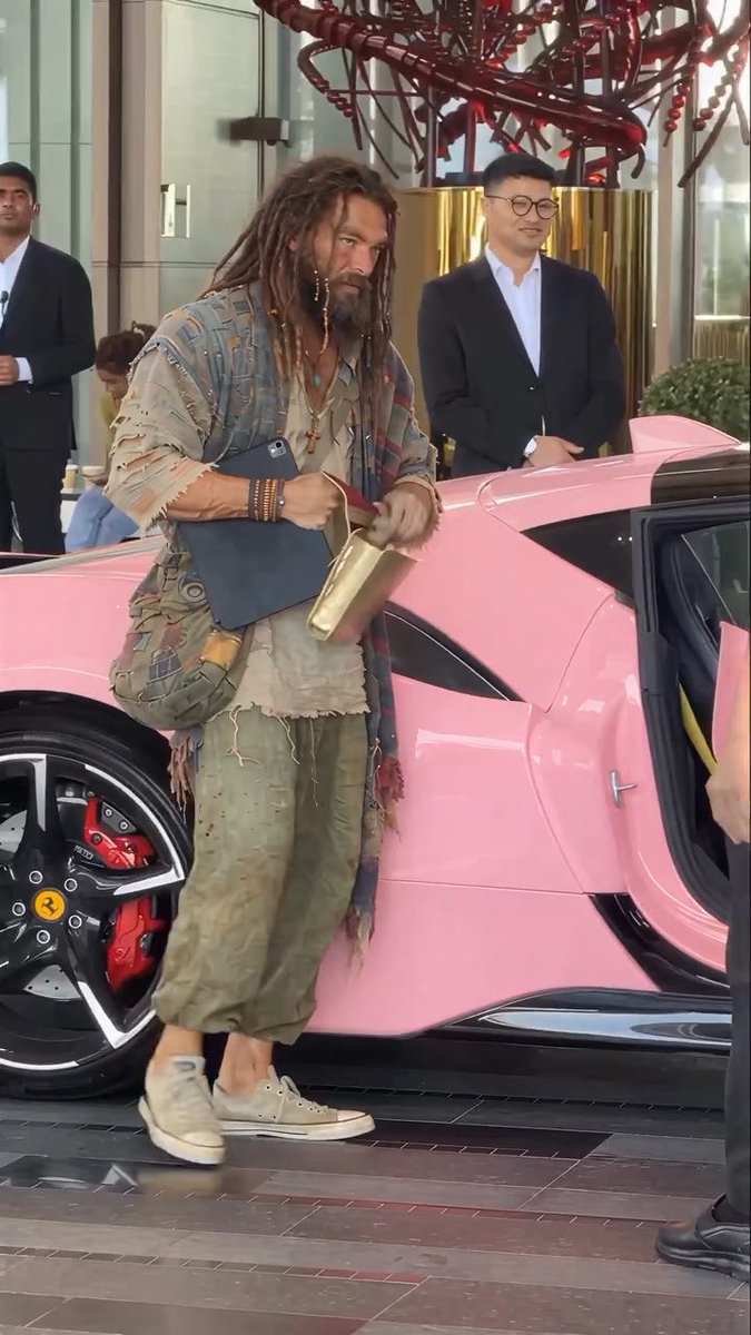 A man with long dreadlocks and a beard, wearing a distressed, layered outfit, exits a bright pink sports car while carrying a clipboard and folders, accompanied by men in suits in the background.