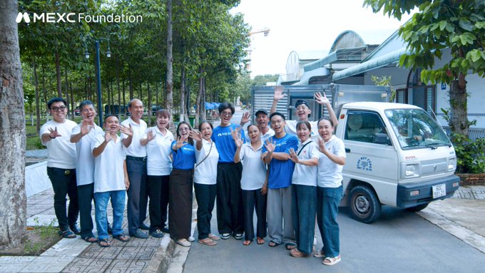 A group photo of people in white and blue shirts posing next to a small white truck with 'MEXC Foundation' logo in the top left corner.