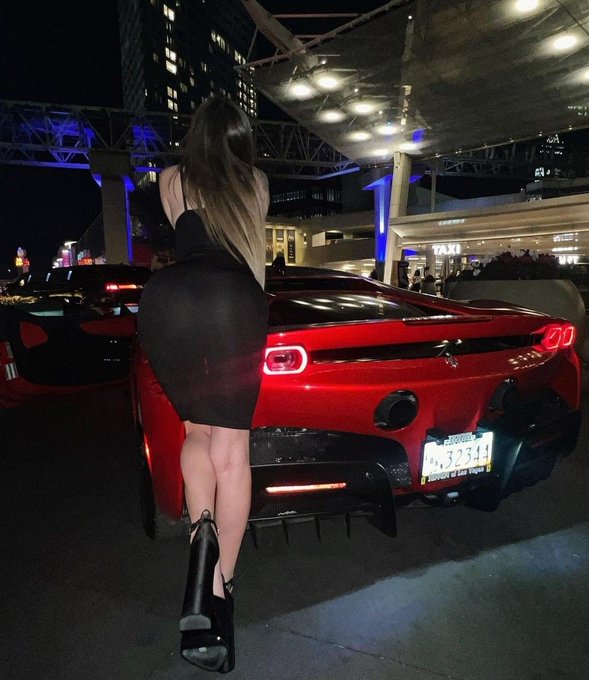 A nighttime photo of a woman in a tight black dress and heels standing in front of a red Ferrari sports car at what appears to be a valet area or car meet.
