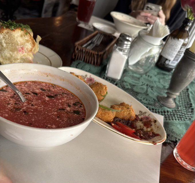 A close-up of a table at a restaurant featuring a bowl of dark red soup, a plate with breaded items, salt and pepper shakers, and a beverage, with partial views of other diners.