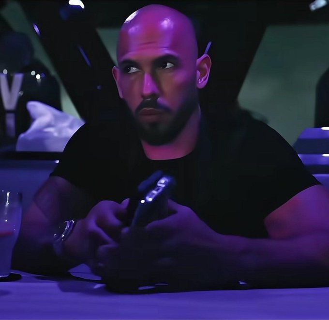 A bald man with a goatee in a black t-shirt holding a phone in a dimly lit setting with purple ambient lighting.