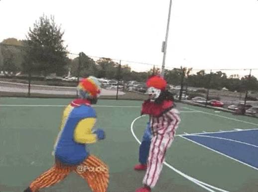 Two people in colorful clown costumes, one blue and yellow and the other red and white stripes, sparring or fighting on an outdoor basketball court.