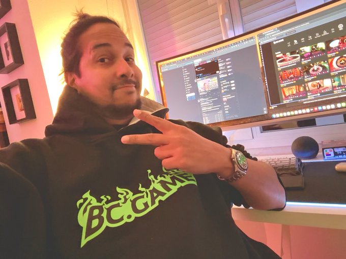 A selfie of a person wearing a black 'BADAM' hoodie, pointing at themselves with a peace sign, sitting in front of dual computer monitors displaying various applications in a warmly lit room.