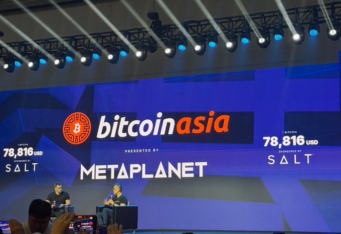 Two people sit on stage at the "Bitcoin Asia" conference presented by "Metaplanet," with the Bitcoin ticker displaying "78,816 USD" alongside a "SALT" sponsorship logo. The image ties to the caption's irony by showing attendees packed into a Bitcoin conference even as Bitcoin trades near $78,816, highlighting the disconnect the post mocks between Bitcoin enthusiasts and their actual holdings.