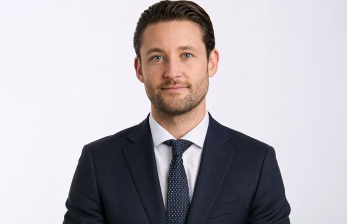 A professional headshot of a man with brown hair wearing a dark suit, white shirt, and a dark patterned tie against a plain white background.