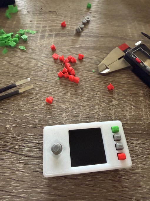 A 3D-printed handheld device with a screen, joystick, and four colored buttons (green, gray, gray, red) sits on a workbench surrounded by loose 3D-printed parts and tools like flush cutters, illustrating a DIY open-source gadget concept. The visible "OFF" and "ON" switch on the cutters and the scattered plastic components reflect the post's discussion about whether to share the design rather than sell assembled units.