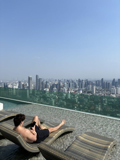 A man relaxes on a lounge chair at a rooftop infinity pool overlooking a sprawling city skyline.