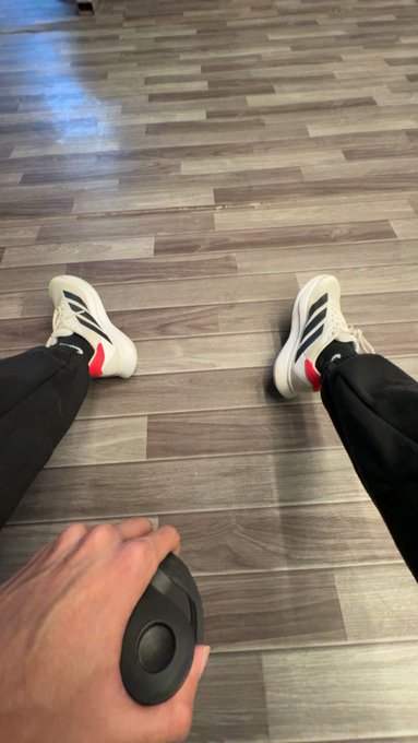 A first-person photo looking down at feet wearing white Adidas sneakers with red and black stripes, black pants, and a hand holding a black computer mouse, standing on a gray wood-grain floor.