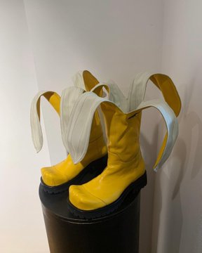 A pair of avant-garde yellow leather boots with silver sculpted heels displayed on a black pedestal in a white gallery space.