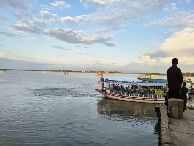 A scenic view of a waterfront with passenger boats docked, a person standing on a pier watching the sunset under a partly cloudy sky.