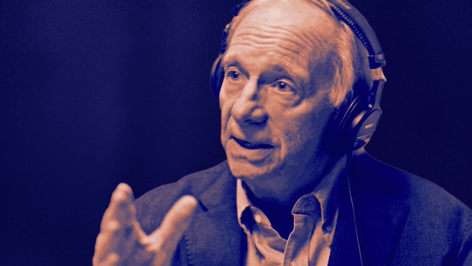 A close-up photo of an older man wearing headphones and a blazer, gesturing with his hand while speaking, set against a dark blue background.