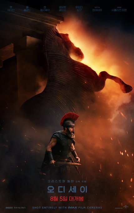 A dramatic movie poster showing a Roman soldier standing before a giant Trojan horse silhouette amid flames and smoke, with Korean text '오디세이' (Odyssey), a release date of '8월 5일 대개봉' (August 5 grand opening), and the note 'SHOT ENTIRELY WITH IMAX FILM CAMERAS'.