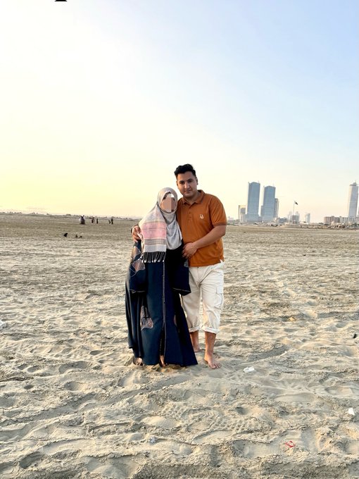 A couple poses together on a sandy beach with a city skyline in the background, sharing a warm embrace as part of their "Good Morning" greeting to followers. The serene seaside setting with skyscrapers in the distance adds a peaceful backdrop to their request for "GM BACK" responses.