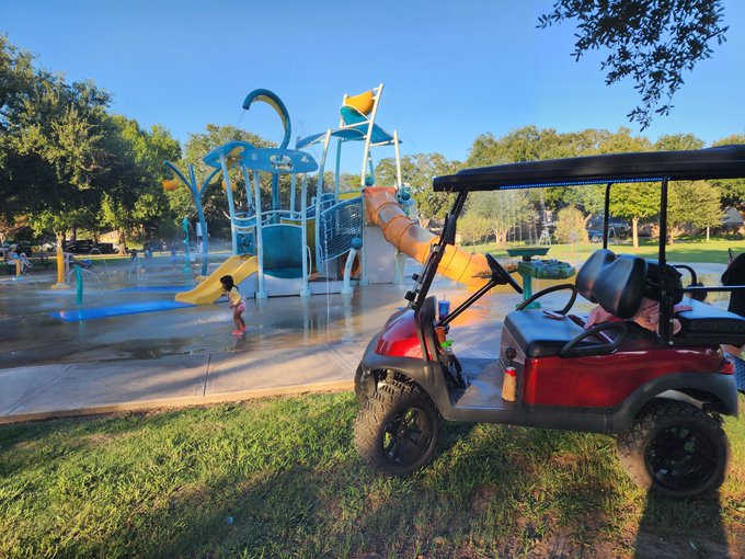 A child plays on a splash pad near a playground while a golf cart sits parked on the grass in the foreground. The image of someone simply enjoying a quiet park moment contrasts with the post's message about playing a different, more patient investment game while others chase quick liquidity.