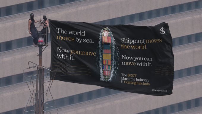 Two workers hang a black banner on a tall building reading 'The world moves by sea. Now you move with it.' and 'Shipping moves the world. Now you can move with it. The $20T Maritime Industry Is Coming Onchain' by Ritual Protocol.