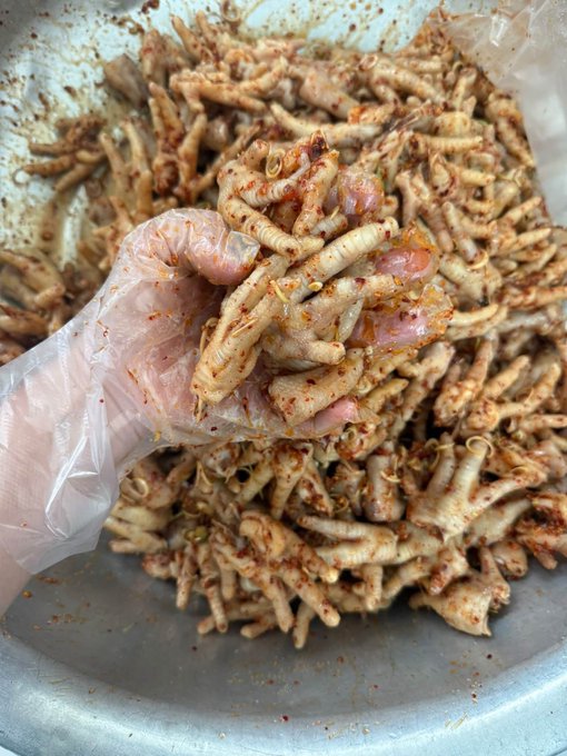 A hand wearing a clear plastic glove holds a handful of cooked chicken feet coated in red chili seasoning over a metal bowl filled with more of the same dish.