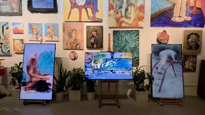 An art gallery space displays various paintings on the walls alongside digital screens showing artworks, with potted plants arranged around the displays.