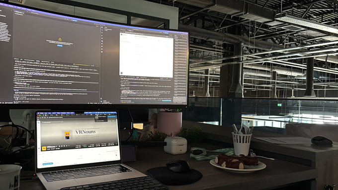 An office desk setup features a laptop displaying 'VRNews' alongside dual monitors showing code and text, with a small plate of food and a computer mouse nearby.