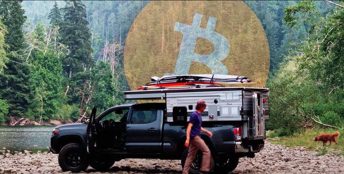 A promotional image showing a person next to a dark pickup truck with a camper shell, a dog, and a large Bitcoin logo overlaid on a forest landscape.