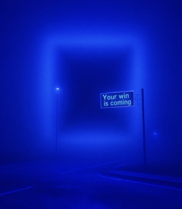 Atmospheric blue-toned image of a foggy road with a glowing neon sign reading "Your win is coming," alongside a single streetlight in the misty background.