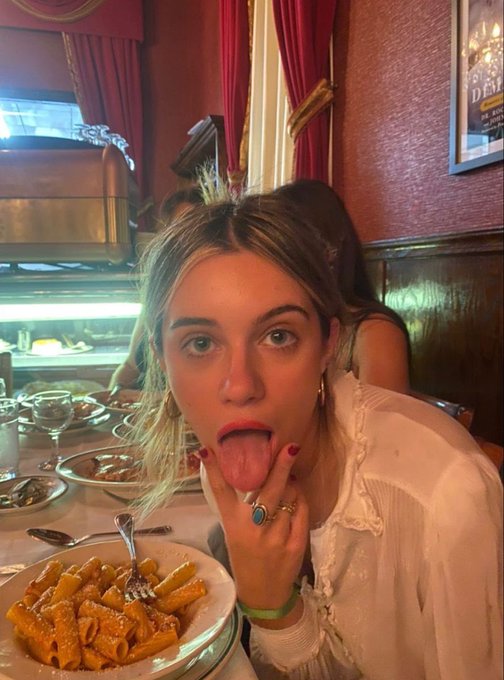 A young blonde woman with red nail polish and a turquoise ring playfully sticks her tongue out at the camera while seated at a restaurant table with a plate of rigatoni pasta in front of her, with a partially visible poster reading "DEM" on the wall behind her.