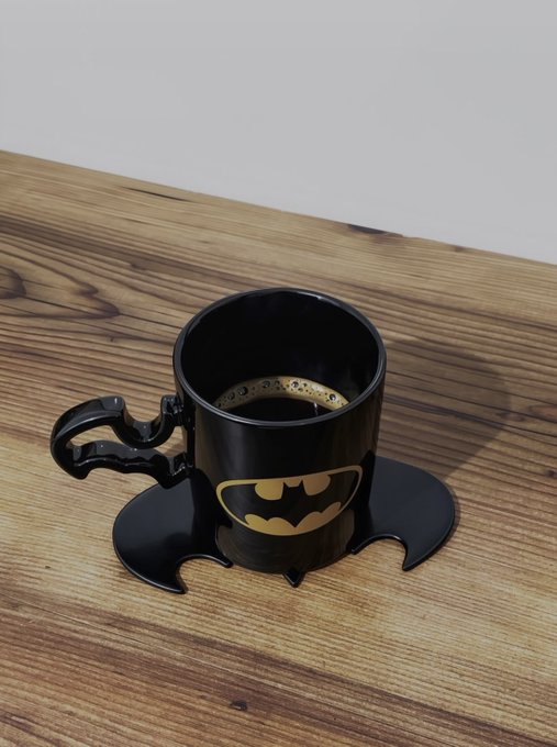 A black Batman-themed ceramic mug sitting on a matching bat-shaped coaster, filled with coffee, on a wooden surface.
