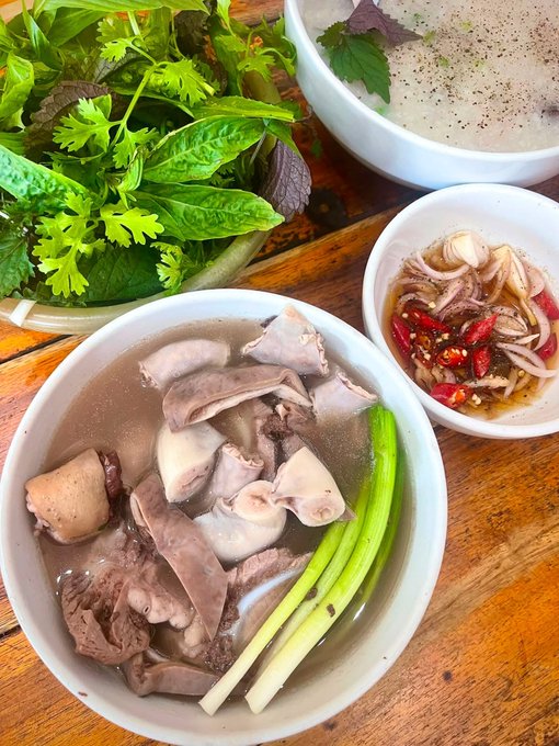A traditional Vietnamese meal featuring a bowl of beef and pork offal pho with green onions, accompanied by fresh herbs, a side of sliced onions with chili, and a dipping sauce on a wooden table.