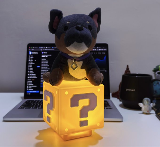 A plush dog toy sitting on a glowing yellow question mark block (resembling a Mario mystery block) on a desk in front of a laptop displaying code and charts.