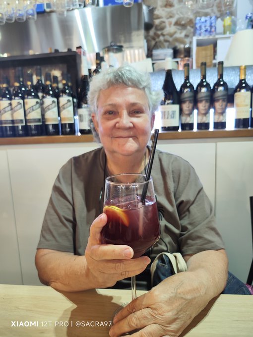 An older woman with short gray hair holding up a glass of red sangria with a straw and orange slice, sitting in front of a bar lined with wine bottles; text reads 'XIAOMI 12T PRO | @SACRA97'.