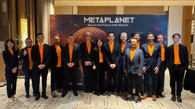 The image features a group of people posing for a photo in front of a large banner that says "Metaplanet". The banner displays the text "Secure the future with Bitcoin" along with an illustration of a planet, possibly representing the concept of Bitcoin. The group consists of men and women standing close together, all wearing suits and ties. They are positioned in front of this banner, creating a visually striking scene that highlights their association with the Metaplanet project.
