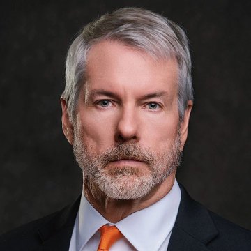 A distinguished, confident-looking man with gray hair and a beard, wearing a dark suit, light blue shirt, and orange tie, gazes directly at the camera in a formal portrait against a dark backdrop. The image visually represents Michael Saylor, the figure celebrated in the post as one of history's greatest investors for his Bitcoin strategy through $MSTR.