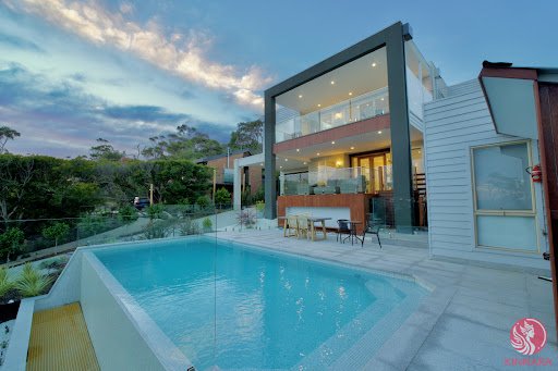 Real estate photograph of a modern multi-story luxury home with a glass-walled upper level, outdoor dining area, and an infinity pool overlooking a forested landscape at dusk, watermarked 'REMIX'.