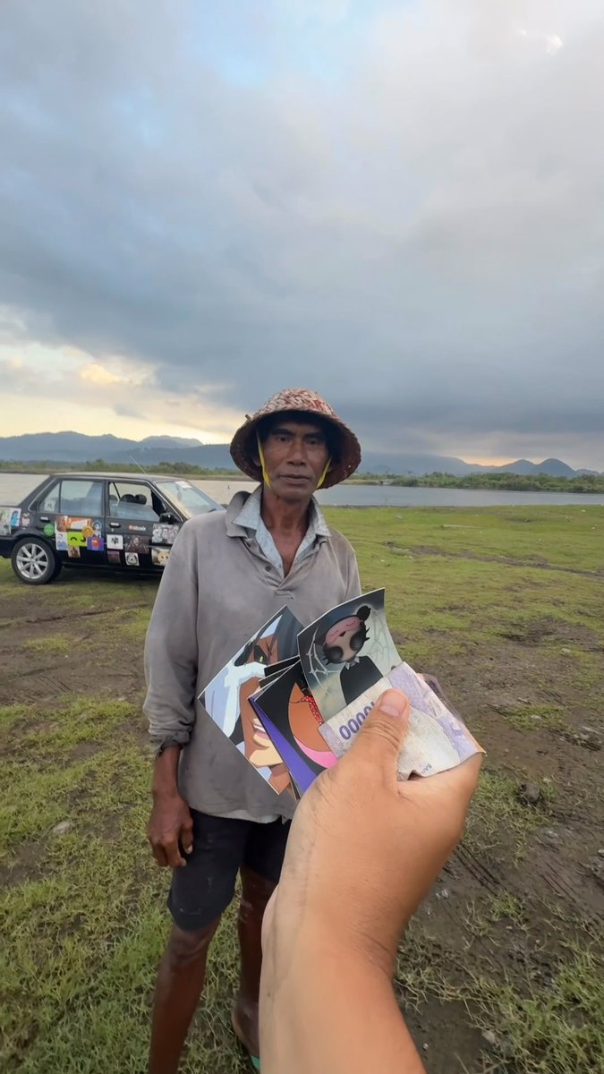 A photograph showing a hand in the foreground holding anime photo cards and what appears to be currency, extended toward an older man wearing a woven hat and a gray shirt who is standing in a grassy field near a decorated car and a body of water.