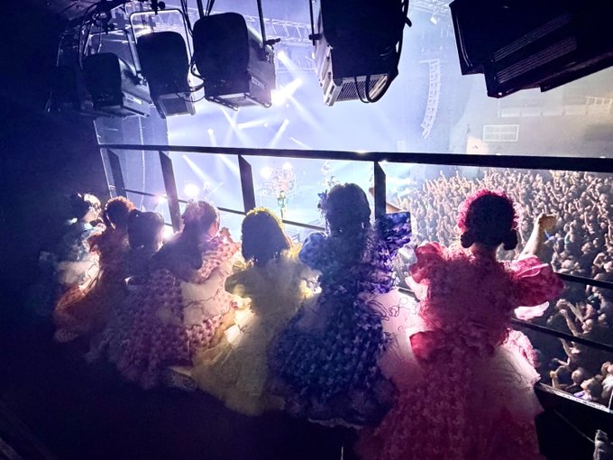 Five performers in elaborate pastel gowns stand on a balcony overlooking a packed arena crowd, with stage lights beaming above, capturing the magical atmosphere of the METALVERSE live show.