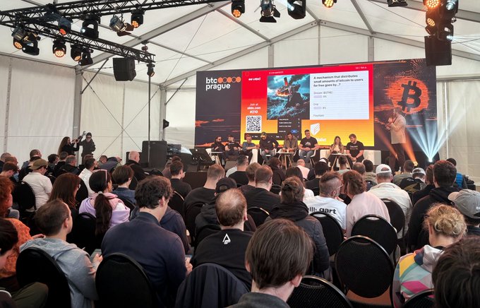 The image shows a large crowd of people gathered inside a tent for an event featuring Bitcoin enthusiasts. The attendees are seated in rows of chairs facing a stage, where a panel discussion or debate about Bitcoin is taking place. A large screen displays information related to the Bitcoin event, and there are multiple speakers on the stage presenting their views on Bitcoin.

The crowd consists of people sitting at various positions within the tent, with some standing near the front row while others are seated further back. The atmosphere appears lively as attendees engage in conversations or listen intently to the presentations.