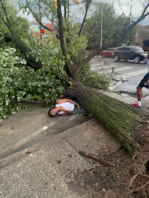 A fallen tree lies across a sidewalk and street after apparently being uprooted, with a person resting underneath the trunk and another person in red and white sneakers standing nearby.