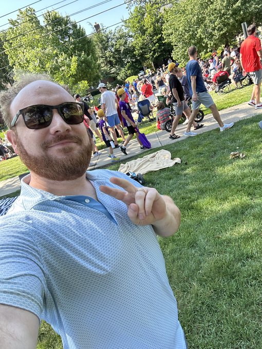 A bearded man wearing sunglasses and a white patterned polo shirt takes a selfie making a peace sign at an outdoor event with people walking in the background and trees lining the area.
