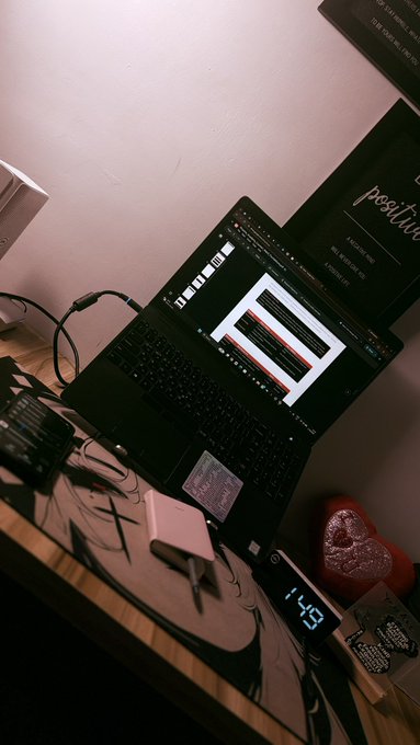 A dimly lit desk setup showing a laptop displaying an audio editing software with a digital clock reading '1:49', surrounded by a pink heart plush and various cables.