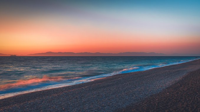 A serene beach landscape at sunset, featuring a gradient sky of orange, pink, and blue hues over calm ocean waters and a pebbly shoreline with distant mountains on the horizon.