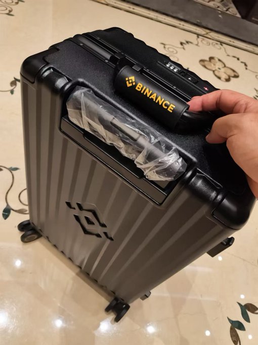 A sleek gray suitcase featuring the Binance logo is displayed on a tiled floor, with a hand resting on its black handle and what appears to be plastic-wrapped accessories in the front compartment. The image showcases the upgraded promotional gift from Binance, highlighting the more subtle branding that addresses earlier complaints about oversized logos.